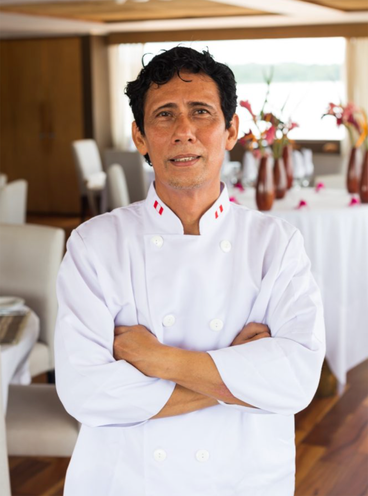 Chef at Delfín Amazon Cruises standing onboard in the dining area wearing a white uniform